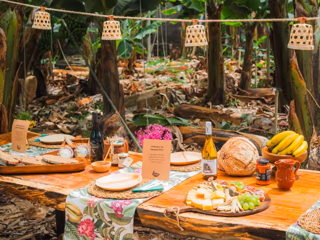Mesa de madera puesta bajo las plataneras con quesos palmeros, pan, mojo, vino y plátanos, iluminada por farolillos de mimbre